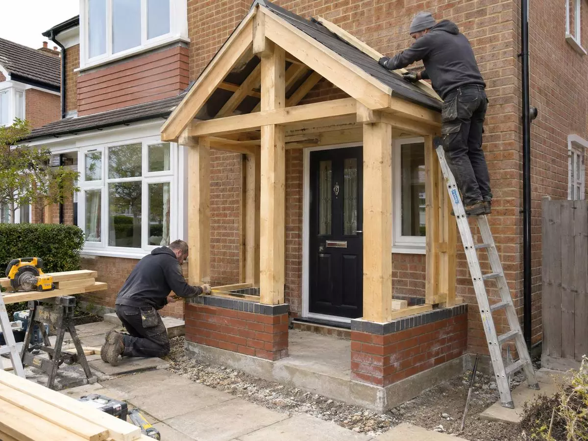 Builders working on a timber-framed porch with a brick base and pitched roof.
